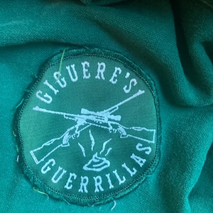 Może przedstawiać: Zielona łatka z białymi szwami i tekstem "Ciguere's Guerrillas" w kółku. W środku łatki znajduje się stylizowany obraz dwóch skrzyżowanych karabinów z celownikiem na górze.