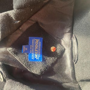 May include: Close-up of a black wool coat with a satin lining. A blue tag reads "PENDLETON WOOLEN MILLS" and "WARM ANTI-TO-BE-A". A gold button is visible. The coat is size 20.