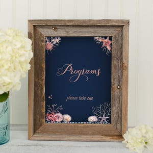 May include: A rustic wooden frame with a navy blue background and pink and white sea life illustrations. The text "Programs" is written in pink script and the text "please take one" is written in white.