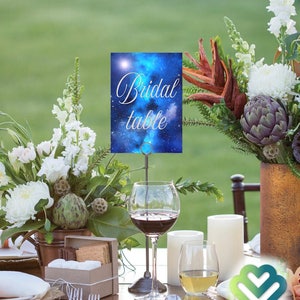 May include: A blue and white galaxy themed table card with the text "Bridal Table" on it. The card is displayed on a metal stand with a glass of red wine and a glass of white wine on a wooden table.