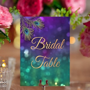 May include: A purple and teal table number card with gold glitter and peacock feathers. The card says "Bridal Table" in gold script.