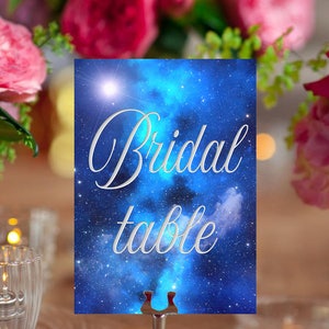 May include: A blue and white galaxy themed table card with the text "Bridal table" in silver lettering. The card is displayed on a silver stand.