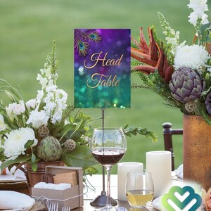 May include: A purple and green table number card with gold lettering that reads "Head Table" and a peacock feather design. The card is on a metal stand and is placed on a table with a floral centerpiece, wine glasses, and candles.