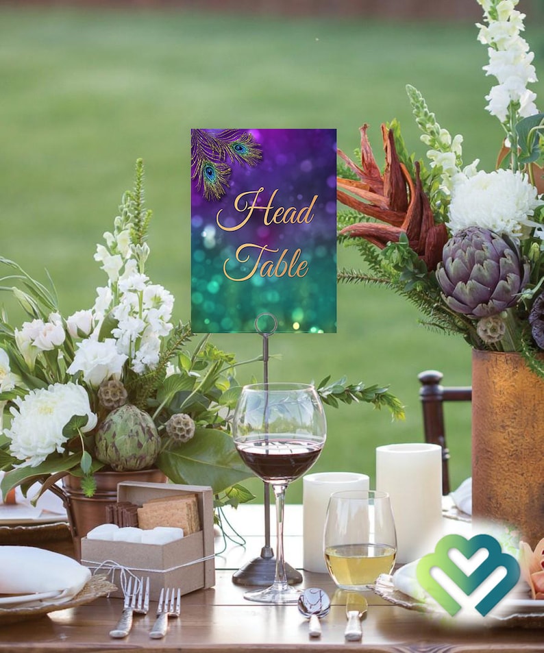 May include: A purple and green table number card with gold lettering that reads "Head Table" and a peacock feather design. The card is on a metal stand and is placed on a table with a floral centerpiece, wine glasses, and candles.