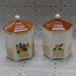 May include: Two white ceramic jars with brown and tan pagoda-style lids. Each jar has a floral design and a decorative, geometric pattern. The jars are on a white floral patterned surface.