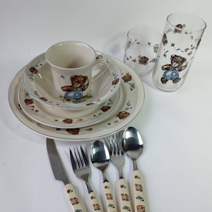 Peut inclure: Un ensemble de vaisselle pour enfants avec un motif d'ours en peluche. L'ensemble comprend un bol, une assiette, une tasse et deux verres. La vaisselle est blanche avec une bordure bleue et représente un ours en peluche portant une salopette. Les couverts sont également décorés avec le motif de l'ours en peluche.
