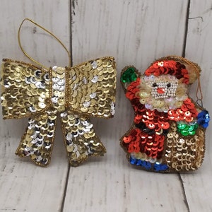May include: Two Christmas ornaments: a gold sequined bow and a Santa Claus figure. The bow is made of gold and silver sequins. The Santa ornament is red, gold, and blue, with a white beaded face and a gold sequined body.