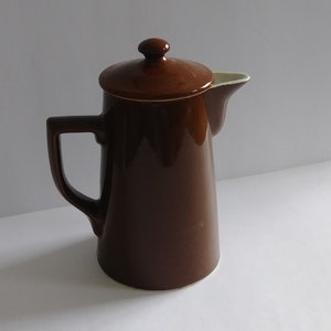 May include: A brown ceramic coffee pot with a lid and handle. The pot has a classic design and a glossy finish. The spout is visible on the right side of the pot. The background is a plain white surface.