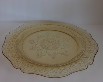 May include: A vintage amber-colored glass serving plate with a decorative, scalloped edge. The plate features an embossed floral and geometric design in the center and along the rim. The plate is round and has a slightly raised edge.