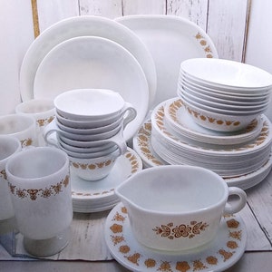 Peut inclure: Un ensemble de vaisselle blanche ornée d'un motif floral doré. L'ensemble comprend des assiettes, des bols, des tasses, des soucoupes et une crémière. La vaisselle est empilée et disposée sur une surface en bois clair.