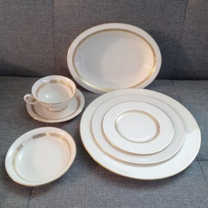 May include: A collection of white porcelain dishware with gold leaf trim. The set includes an oval platter, a teacup and saucer, a small oval dish, and a stack of plates. The gold trim features a repeating leaf pattern.