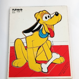 May include: A vintage Playskool Pluto 3D puzzle. The yellow dog with black ears and a red tongue is holding a white bone. The puzzle is on a red and white background. The puzzle is made in the USA.