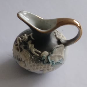 May include: A small, dark grey ceramic jug with a gold handle and rim. The jug features a raised relief design in cream and light blue, with a curved spout. The handle is a rounded loop.