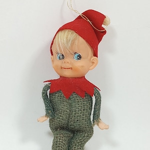 May include: A vintage Christmas elf ornament. The elf has a plastic face with blue eyes and blonde hair. It wears a red felt hat and collar, and a green burlap suit with red feet. A gold string is attached for hanging.