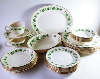 Könnte beinhalten: Ein komplettes Vintage-Tafelservice aus weißem Porzellan mit einem grünen Efeublattmuster und Goldrand. Das Set umfasst Teller, Schalen, Tassen, Untertassen und Servierschalen, ideal für ein formelles Abendessen.