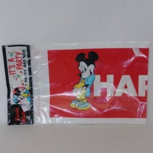 May include: A clear plastic package containing a red banner with the word "HAPPY" in white and a Mickey Mouse graphic. A smaller black and white banner with the text "IT'S A PARTY" is also visible.