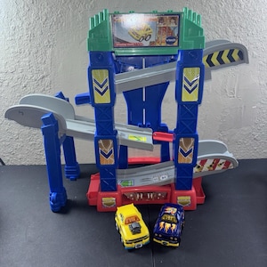 May include: A VTech toy playset featuring a multi-level blue and green plastic track structure with gray ramps, yellow and blue chevron accents, and a top sign displaying a yellow car. Two small toy cars, one yellow and one dark blue, sit in front.