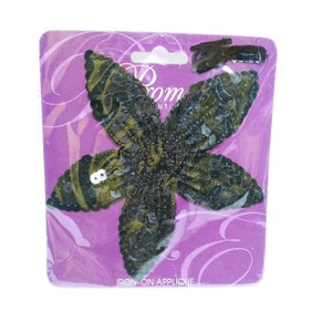 May include: An iron-on applique in the shape of a flower, with olive green and black sequins. The applique is on a purple backing with the words "Prom Essentials" and "Iron-On Applique".