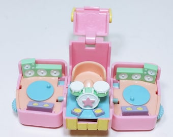 May include: A pink, vintage toy playset featuring a DJ booth and drum set. The playset has a light pink exterior with pastel blue, green, and yellow accents. The drum set has a star design. The toy is open, revealing the interior.