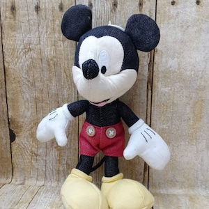 K&ouml;nnte beinhalten: Ein Mickey Mouse Stofftier mit schwarzen denimartigen Ohren, Kopf und Armen. Das Spielzeug hat ein wei&szlig;es Gesicht, H&auml;nde und gelbe Schuhe. Es tr&auml;gt rote Shorts mit zwei braunen Kn&ouml;pfen.
