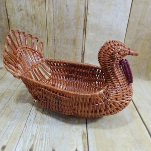 May include: A brown wicker basket shaped like a turkey. The basket has a woven design with a tail fanned out at the back and a turkey head with a red wattle. The basket is designed to hold items.