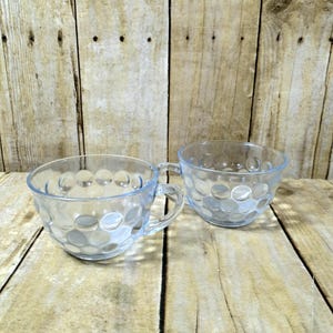 May include: Two clear glass teacups with handles. The cups have a pattern of raised, circular dots. The cups are on a wooden surface with a light brown color.