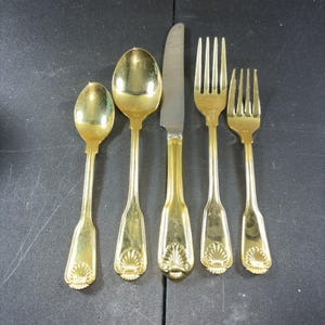 May include: A set of gold-colored flatware including two spoons, a knife, and two forks. The handles have a decorative shell design. The knife has a silver-colored blade. The flatware is arranged on a dark surface.