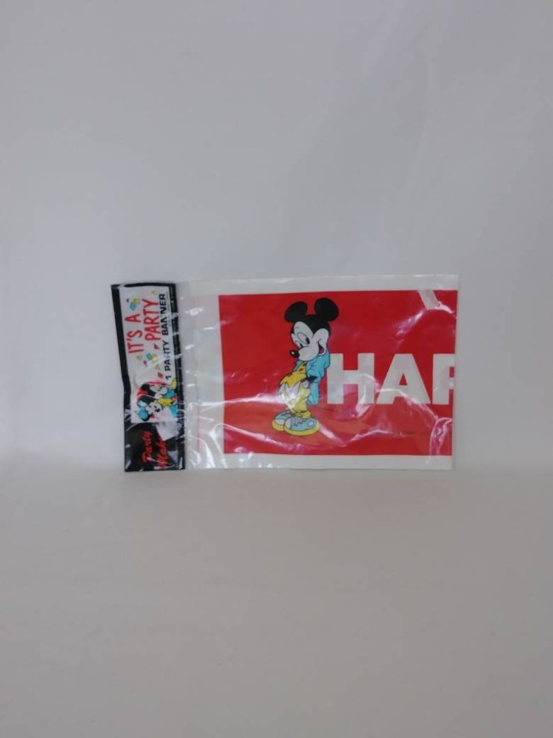 May include: A clear plastic package containing a red banner with the word "HAPPY" in white and a Mickey Mouse graphic. A smaller black and white banner with the text "IT'S A PARTY" is also visible.