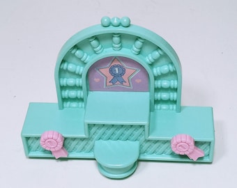 May include: A light teal plastic toy stage with a star-shaped emblem that has a number 1 on it. The stage has pink ribbons on the sides and a decorative arch with small spheres on top.