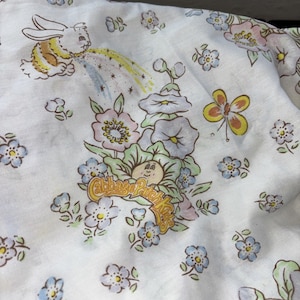 May include: White fabric with a repeating pattern of flowers, butterflies, and a bunny. The fabric features a central illustration of a Cabbage Patch Kid surrounded by flowers. The Cabbage Patch Kids logo is visible.