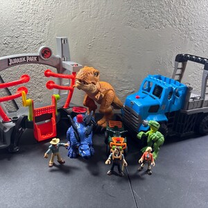 May include: A Jurassic Park playset featuring a gray gate structure with red accents, a brown Tyrannosaurus Rex, a blue Triceratops, a blue transport truck, a small green all-terrain vehicle, a green raptor, and three small human action figures.