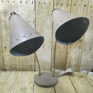 May include: A pair of vintage desk lamps with taupe conical shades and star cutouts. The lamps have flexible, brass-colored gooseneck arms and a matching taupe base. A white power cord with a pink rubber band is visible.