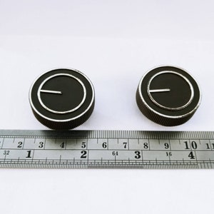 May include: Two black control knobs with silver accents and a ridged edge. Each knob has a white line indicator. A ruler is placed below the knobs, showing measurements in inches.
