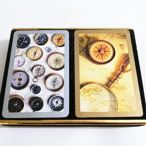 May include: A playing card set featuring compass designs. One card displays various compasses on a map background, while the other shows a compass, a map, and a magnifying glass. The cards are in a black and gold case.