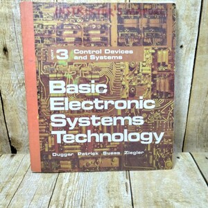 May include: A vintage textbook titled "Basic Electronic Systems Technology" with a cover featuring a circuit board design. The book has a red spine and the text "Unit 3 Control Devices and Systems" is visible. The book is on a wooden surface.