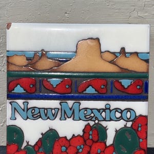 May include: A square ceramic tile featuring a desert landscape with tan mesas under a white sky. A decorative band of red and blue shapes sits above the text New Mexico in light blue letters. The bottom shows green cacti and red flowers.