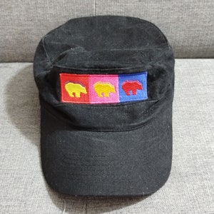 May include: Black military-style cap with a flat peak. The cap has a rectangular patch with three colourful bear designs: yellow, yellow, and red. The bears are embroidered on red, pink, and blue backgrounds.