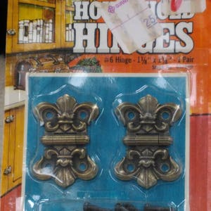 May include: A package of two ornate brass hinges with a fleur-de-lis design. The hinges are 1 1/8 inches by 1 1/2 inches and come with screws. The package is labeled "Household Hinges" and "#6 Hinge - 1 1/8" x 1 1/2" - 1 Pair".