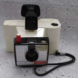 May include: Vintage Polaroid Swinger Land Camera with a black and silver body, red button, and black lens. A black flash attachment and a cream-colored case are behind the camera. The camera has a black strap.
