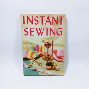 May include: A vintage book titled "Instant Sewing" with red text. The cover features an hourglass, sewing tools, and fabric. A gold hourglass, scissors, thimble, and other sewing notions are displayed.