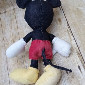 K&ouml;nnte beinhalten: Ein Stofftier von Mickey Mouse, von hinten gesehen. Die Puppe hat einen schwarzen Kopf und Ohren, rote Shorts, wei&szlig;e Handschuhe und gelbe Schuhe. Der Schwanz ist schwarz. Die Puppe ist aus Stoff und befindet sich auf einer Holzoberfl&auml;che.