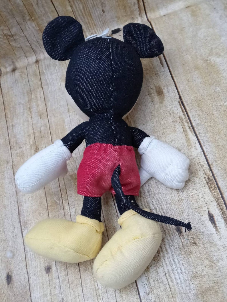 K&ouml;nnte beinhalten: Ein Stofftier von Mickey Mouse, von hinten gesehen. Die Puppe hat einen schwarzen Kopf und Ohren, rote Shorts, wei&szlig;e Handschuhe und gelbe Schuhe. Der Schwanz ist schwarz. Die Puppe ist aus Stoff und befindet sich auf einer Holzoberfl&auml;che.