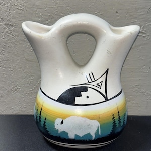 May include: A cream-colored ceramic wedding vase with two spouts and a central bridge handle. The body features a painted scene of a white buffalo standing in a forest against a yellow and blue gradient sky, with a black geometric design above.
