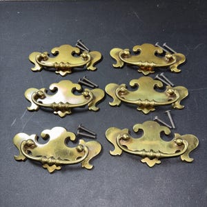 May include: Six antique brass drawer pulls with decorative scrollwork and matching screws. The pulls have a curved handle and ornate backplate. The hardware is a warm gold color, perfect for vintage furniture restoration projects.