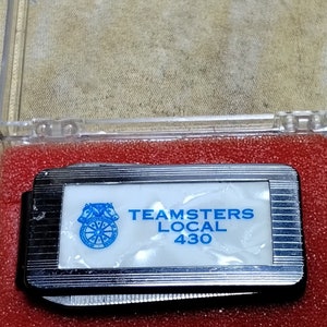 May include: A silver pocket knife with a white pearlescent inlay featuring the text "TEAMSTERS LOCAL 430" in blue. The knife is displayed in a clear plastic case with a red foam insert.