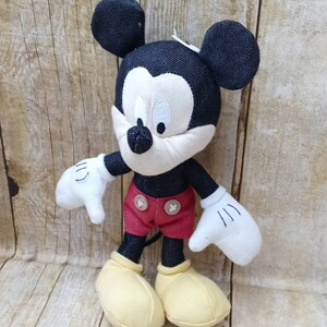 K&ouml;nnte beinhalten: Pl&uuml;schtier Mickey Mouse mit schwarzem Kopf und Ohren, wei&szlig;em Gesicht und wei&szlig;en Handschuhen. Die Puppe tr&auml;gt rote Shorts mit zwei braunen Kn&ouml;pfen und gelbe Schuhe. Der Hintergrund ist eine hellbraune Holzoberfl&auml;che.