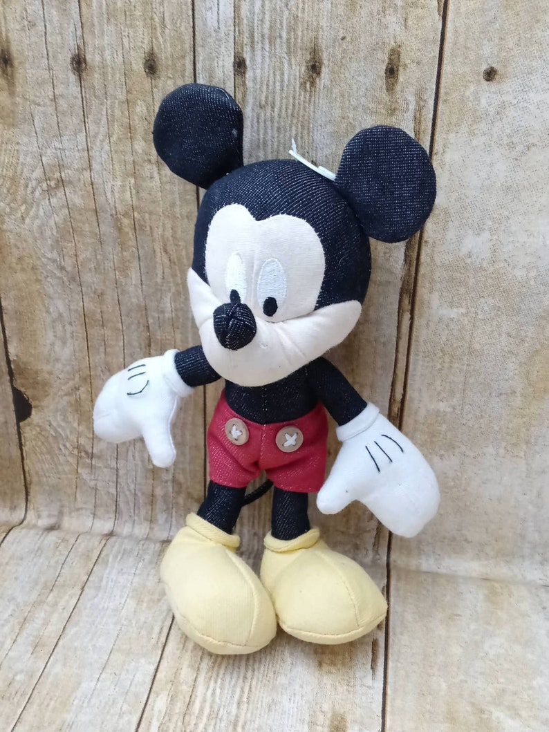 K&ouml;nnte beinhalten: Pl&uuml;schtier Mickey Mouse mit schwarzem Kopf und Ohren, wei&szlig;em Gesicht und wei&szlig;en Handschuhen. Die Puppe tr&auml;gt rote Shorts mit zwei braunen Kn&ouml;pfen und gelbe Schuhe. Der Hintergrund ist eine hellbraune Holzoberfl&auml;che.