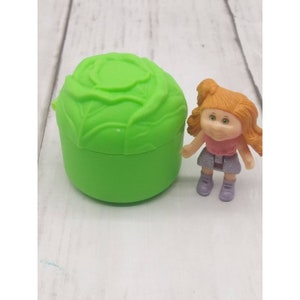 May include: A green plastic cabbage-shaped container with a matching lid, next to a small doll. The doll has orange hair, a pink top, a sparkly silver skirt, and purple shoes. The container is a bright, solid green color.
