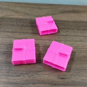 May include: Three bright pink miniature toy suitcases are arranged on a wooden surface. Each suitcase has a rectangular shape with a strap detail. The suitcases are made of plastic.
