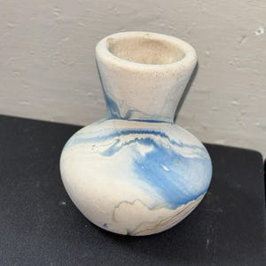 May include: A small, handcrafted vase with a rounded body and a narrow neck. The vase is primarily white with blue and gray marbling around the body. The opening is a light beige color.
